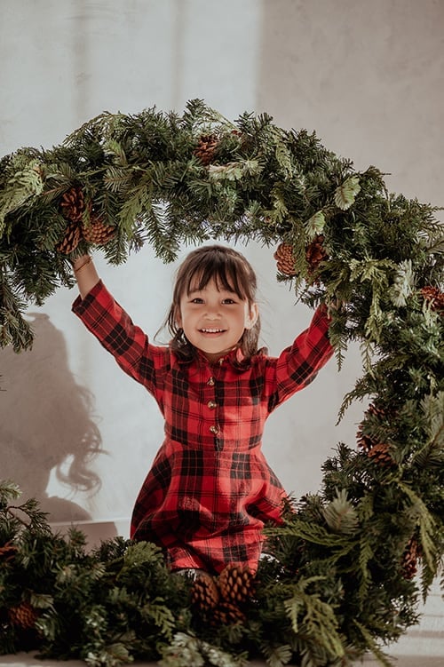 Smiling child in red plaid holding a large evergreen wreath